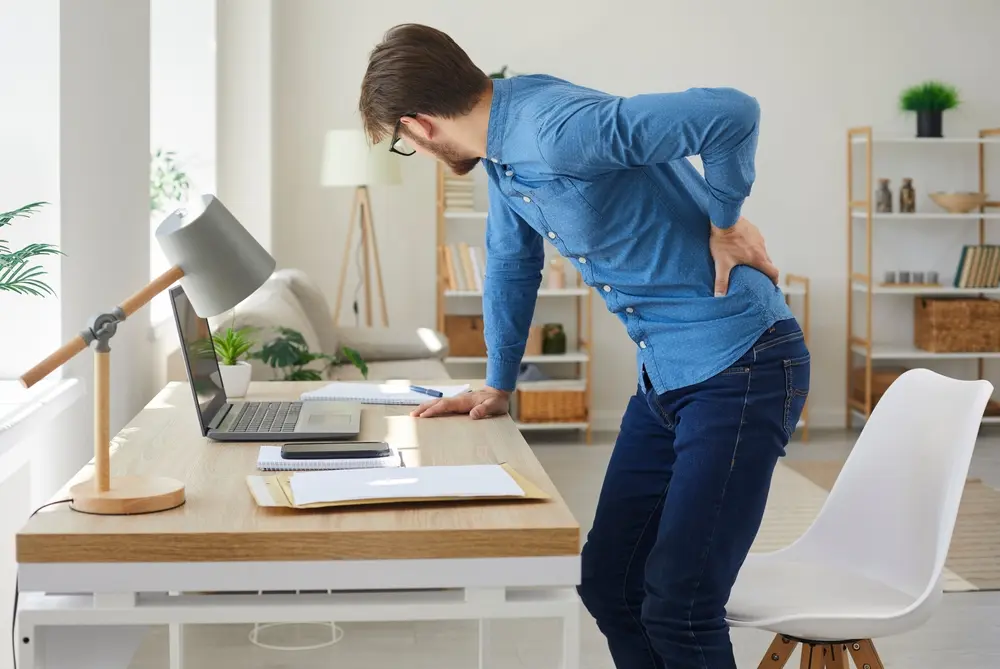 Stressed office worker standing up with intense lower back pain after long sedentary computer work, showing possible lumbar herniated disc or pinched nerve